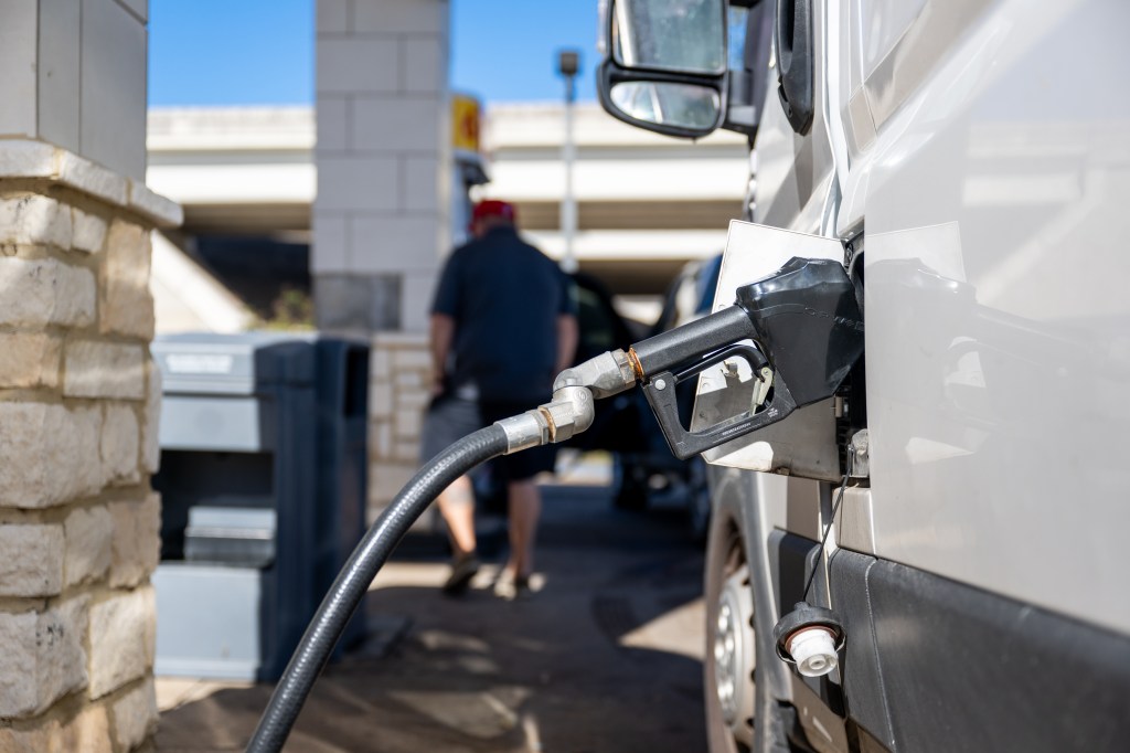 A gas pump nozzle in a vehicle, with a man blurred in the background.
