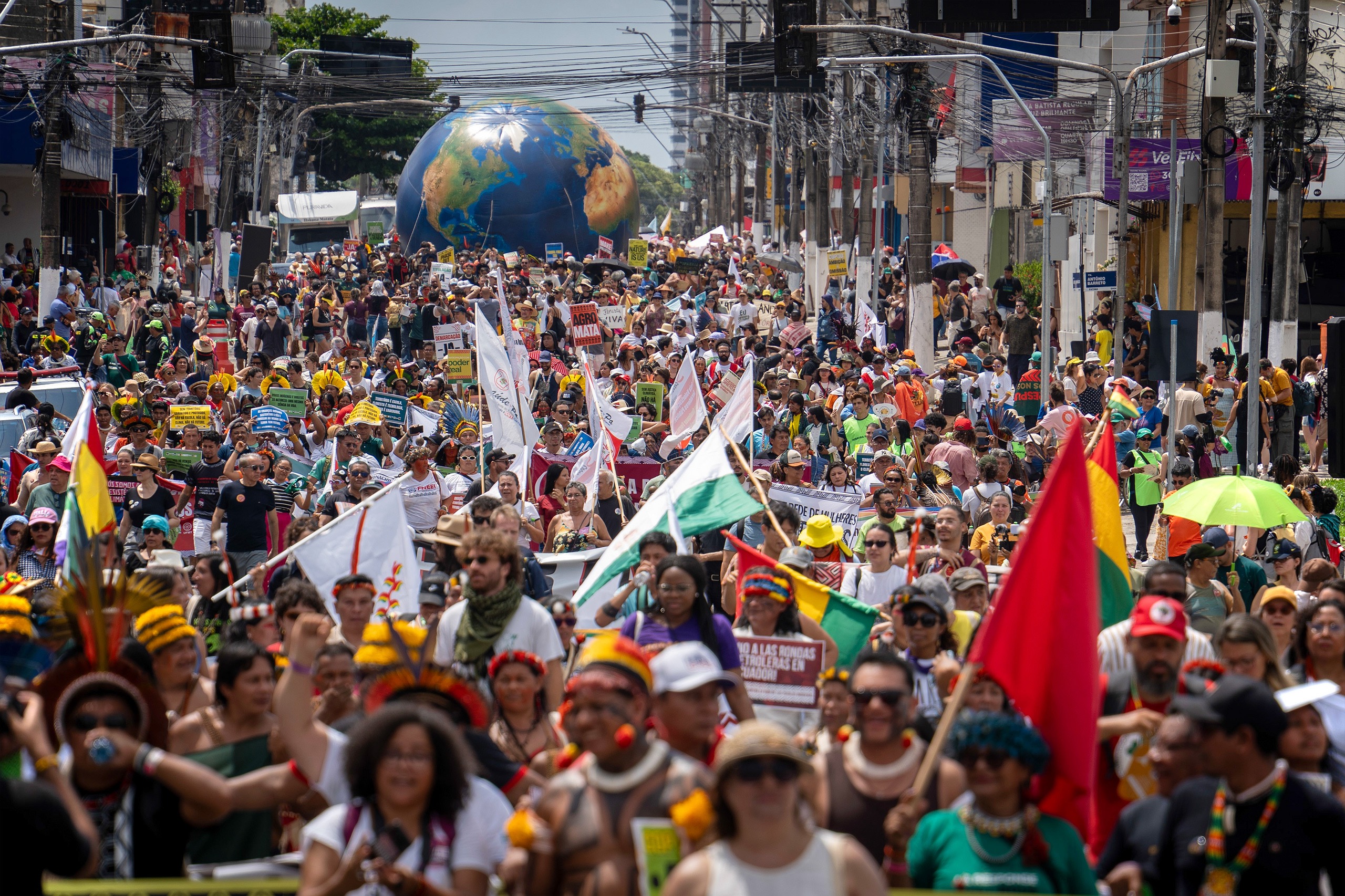 A crowd marches in a city street, holding banners and flags, with a large globe balloon in the background