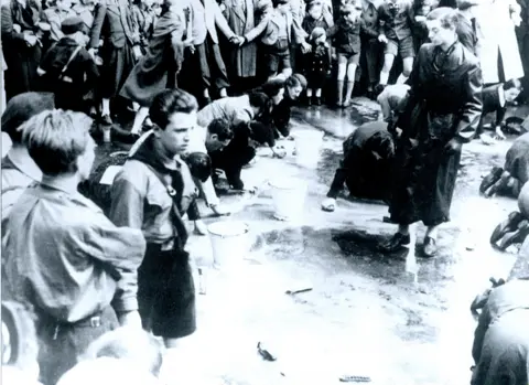 Universal History Archive via Getty Images Black and white photo of Jewish men and women scrubbing a street with sponges while people stand around them