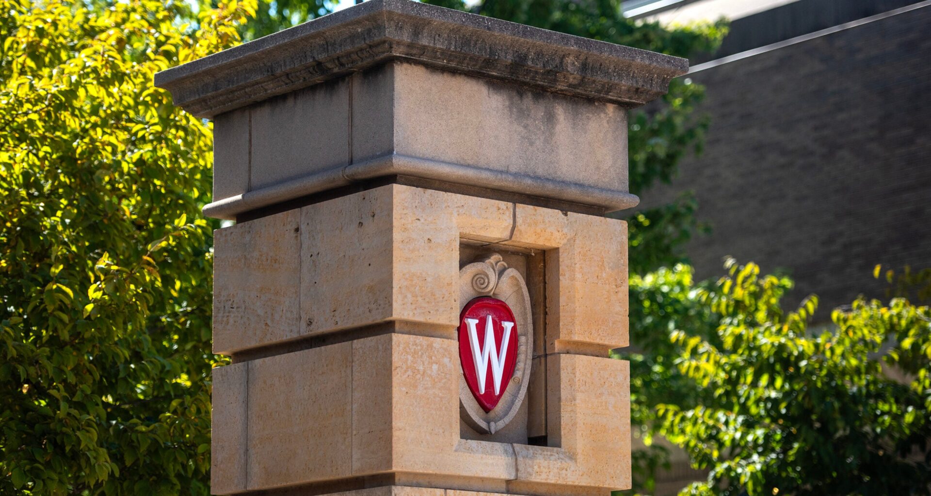A stone pillar with the University of Wisconsin crest featuring a red W is shown against a background of trees and a building.