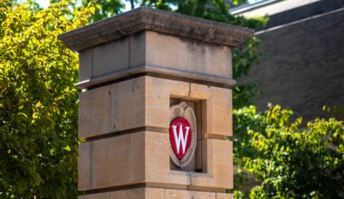 A stone pillar with the University of Wisconsin crest featuring a red W is shown against a background of trees and a building.
