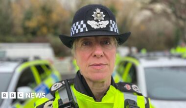 A headshot of a woman wearing police gear. She has a yellow high-visibility jacket on and a black hat. She is looking directly at the camera.
