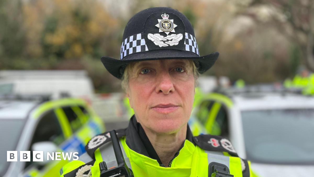 A headshot of a woman wearing police gear. She has a yellow high-visibility jacket on and a black hat. She is looking directly at the camera.