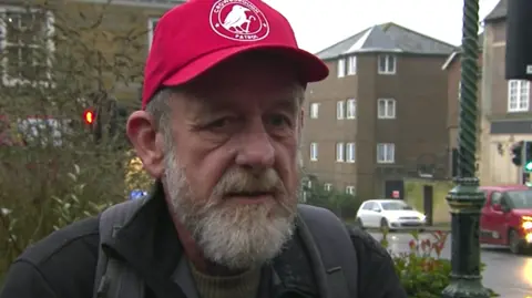 BBC / Piers Hopkirk A man with grey hair and beard is wearing a red baseball cap. He is wearing a black jacket. 