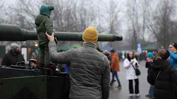 Mies pitää panssarivaunun päällä seisovan lapsen kädestä. Taustalla muita ihmisiä.