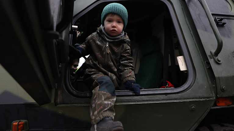 Pieni poika tulossa pois sotilasajoneuvon kyydistä. Hän katsoo kameraan.