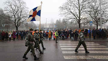 Sotilaat marssivat Suomen lipun kanssa kadulla. Kadun varrella ihmisiä.