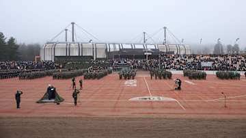 Sotilasjoukkoja sekä kuvaajia pesäpallokentällä. Taustalla Kouvolan jäähalli ja katsomoissa ihmisiä.