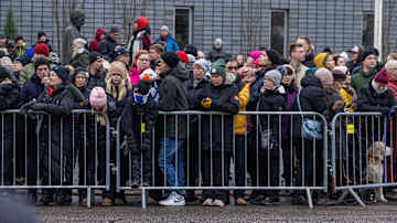 ihmisiä kadun ja rakennuksen välissä metallisten kaiteiden takana.