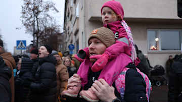 Pinkkeihin vaatteisiin pukeutunut lapsi aikuisen hartioilla väkijoukon keskellä kadun varrella. 
