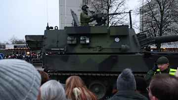 Sotilas seisomassa panssarivaunussa kadulla. Kadun varrella joukko ihmisiä.