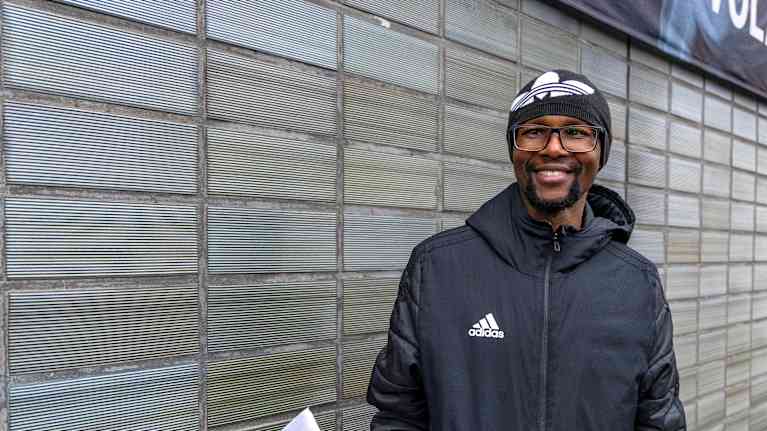 Mies seisoo pipo päässä rakennuksen edessä ja hymyilee Suomen lippu kädessä.