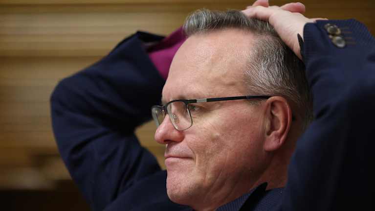 A man with grey hair wearing a dark-blue jacket and purple shirt leans back with his hands behind his head.