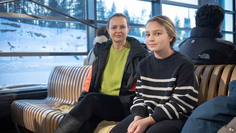 A woman and 10-year-old girl sit on an airport bench, with a snowy scene seen through large windows in the background.