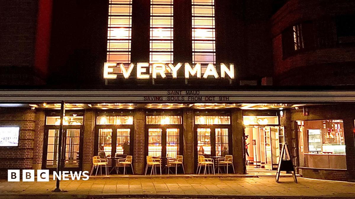 The facade of an art deco-style cinema building at night. There are five patio glass doors with tables and chairs in front. The far right is the entrance to which the doors are wide open and a person can be seen walking inside. Above the doors is a lit LED sign stating: EVERYMAN.