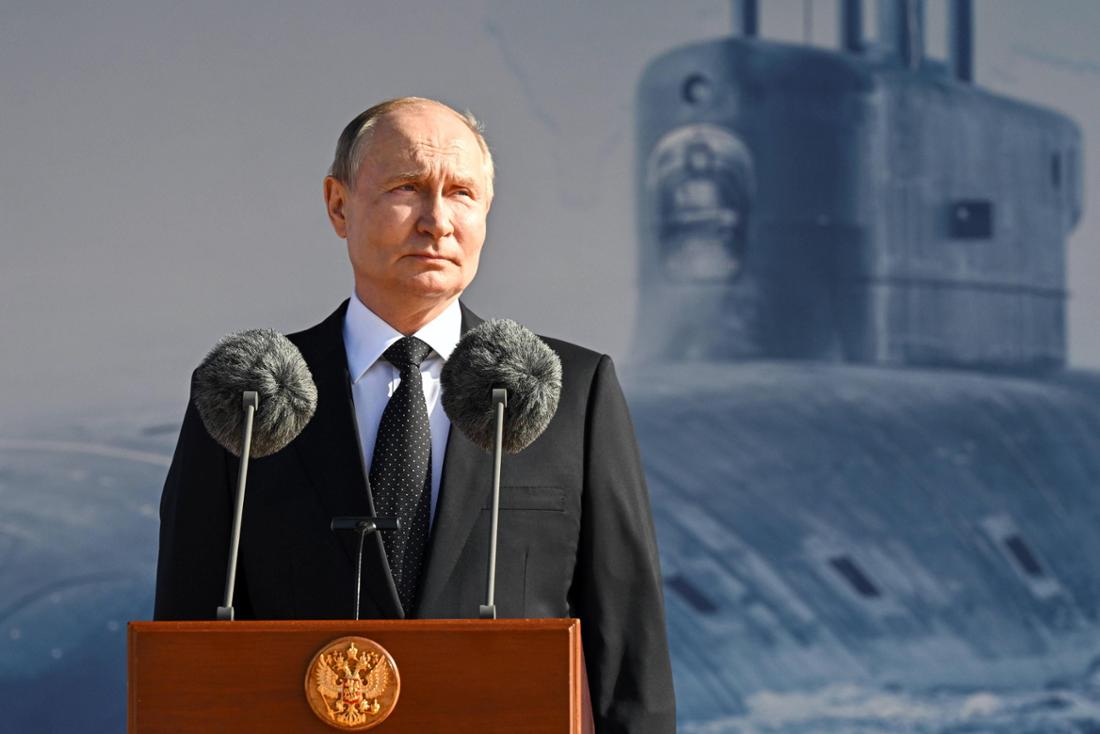 Wladimir Putin vor einem Podium, im Hintergrund einen Fotowand mit dem Druck eines U-Boots.