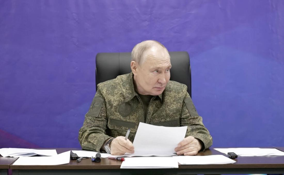 Wladimir Putin sitzt in Camouflage-Uniform an einem Tisch und hält Dokumente und einen Stift in Händen.