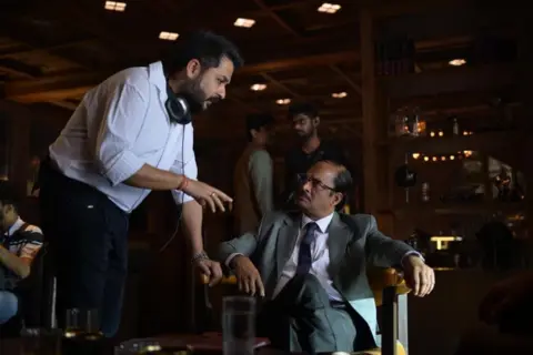 Jio Studios Aditya Dhar, who is dressed in a white shirt and black trousers, leans and talks to R Madhavn, who is wearing glasses and a grey suit with a blue tie, on the sets of Dhurandhar 