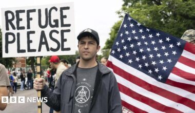 Afrikaans farmer wearing a cap and black jacket, holding a placard written "Refuge Please" behind a US flag