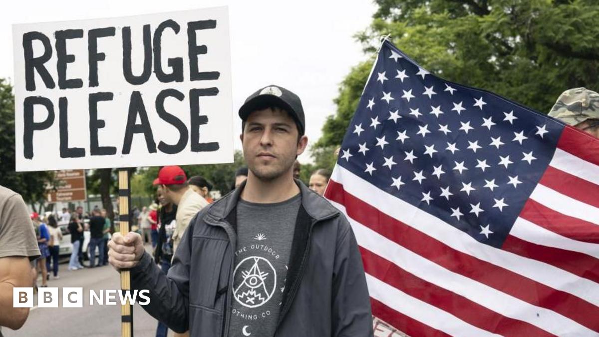 Afrikaans farmer wearing a cap and black jacket, holding a placard written "Refuge Please" behind a US flag