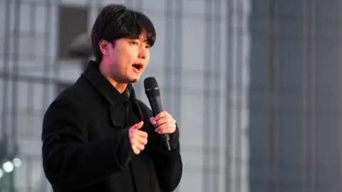 BBC/Hosu Lee A man with black hair and wearing a black speaks into a microphone