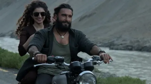 Jio Studios A scene from Dhurandhar movie in which actor Ranveer Singh, wearing an unbuttoned shirt over a vest, and sporting a beard, is riding a motorbike, next to a river. Sitting on the pillion is actress Sara Arjun, wearing a black kurta and sporting aviator sunglasses