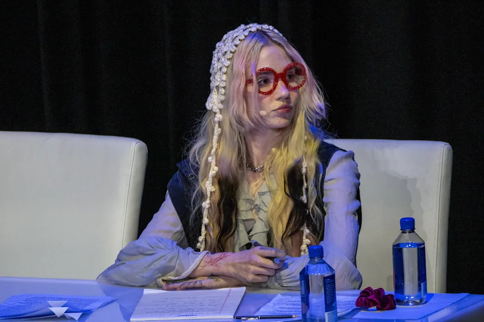 Grimes sits at a table in a formal setting, wearing red glasses and decorative headwear, with papers and water bottles on the table