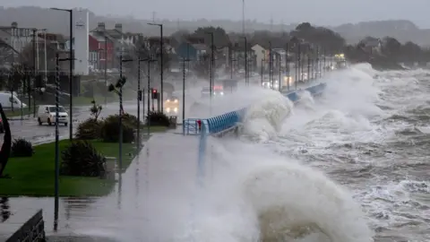 Stephen Henderson Large waves are seen crashing against the seawall in Carrickfergus. A number of cars and a lorry are travelling along the road.