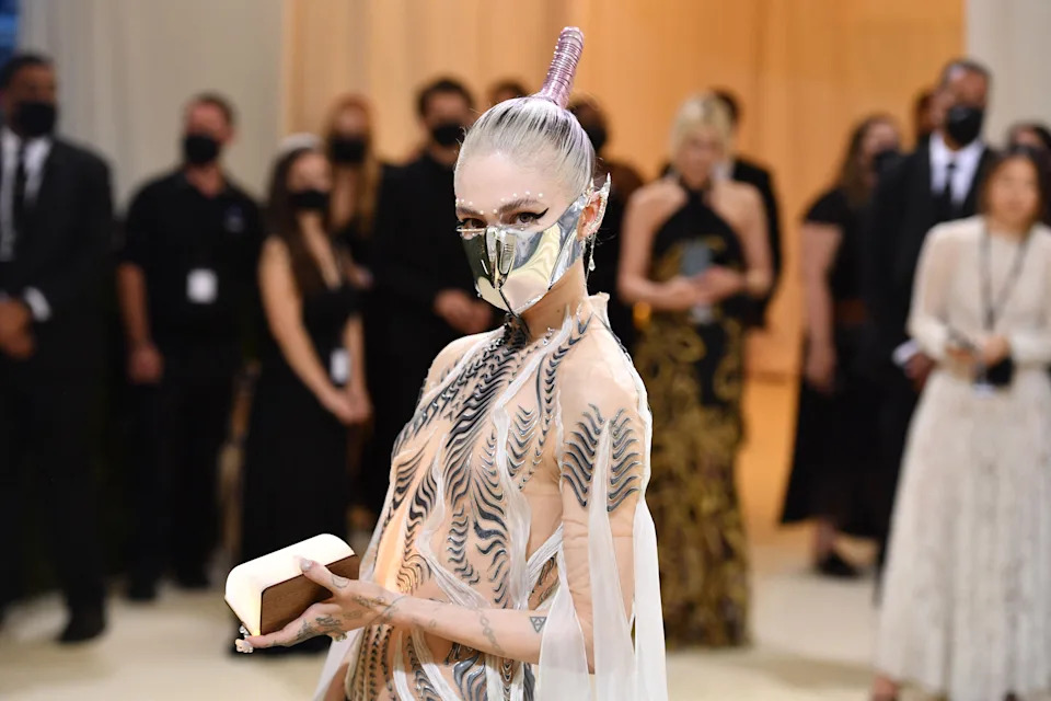 Grimes on a red carpet wearing a futuristic gown with face paint and holding a book, surrounded by masked attendees