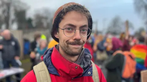 Tomos Hawksworth is a younger man with glasses and neatly trimmed beard. He is wearing a brown hat and red jacket