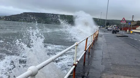 BBC Waves crashing against a sea wall in a village. There are cars travelling down the road. 