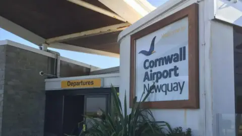 Cornwall Airport Newquay entrance with sign on a curved wall. Automatic doors are visible with a yellow sign above which has the word 'Departures' on it.