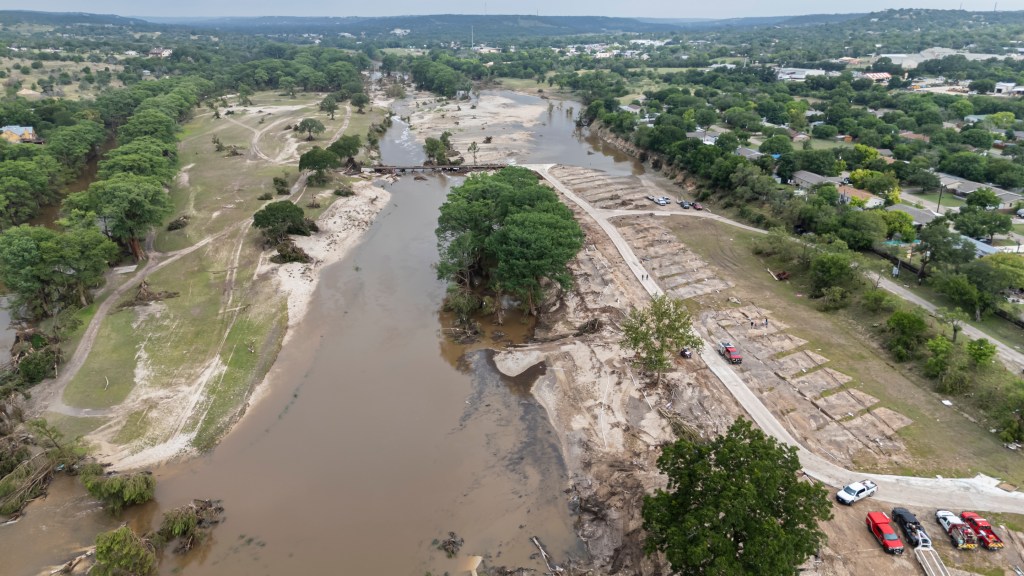 Aerial view of the Blue Oak RV park in Kerrville, TX after floods, showing widespread damage with a muddy river, displaced earth, and vehicles.