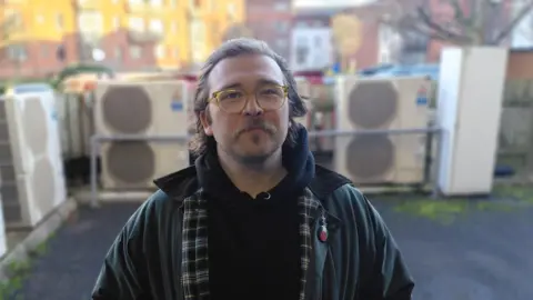 JR McCormick A man with brown hair looks at the camera. He is wearing a jacket and a hoodie, as well as glasses. He is standing outside. There are red-brick buildings in the background. 