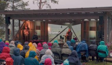 En musikalsk opptreden finner sted utendørs, der en gruppe mennesker sitter på stoler. To utøvere er plassert på en scene, hvor den ene spiller saksofon og den andre står og synger. Publikum, kledd i ulike regnjakker, ser mot scenen, mens omgivelsene er preget av grå skyer og naturlig vegetasjon. I bakgrunnen kan man skimte en utsikt over landskapet. (Bildebeskrivelsen er laget av en KI-tjeneste)