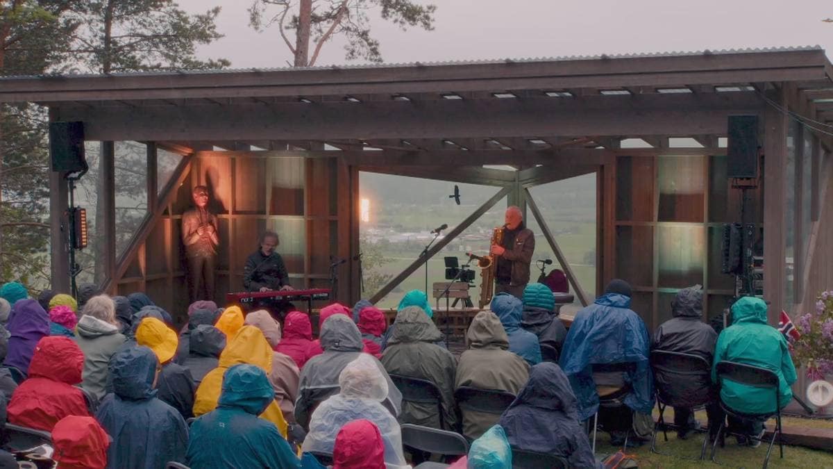 En musikalsk opptreden finner sted utendørs, der en gruppe mennesker sitter på stoler. To utøvere er plassert på en scene, hvor den ene spiller saksofon og den andre står og synger. Publikum, kledd i ulike regnjakker, ser mot scenen, mens omgivelsene er preget av grå skyer og naturlig vegetasjon. I bakgrunnen kan man skimte en utsikt over landskapet. (Bildebeskrivelsen er laget av en KI-tjeneste)