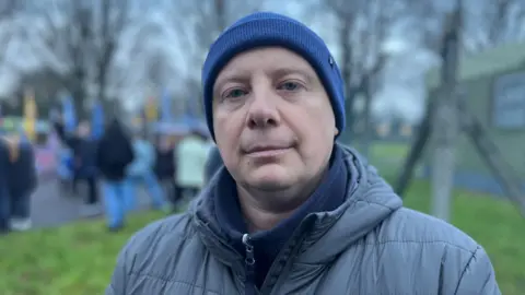 Kamil Frankowski is standing outside with people in the background. He is wearing a blue hat and grey jacket.