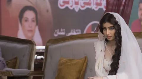 Jio Studios A scene from the movie in which actor Sara Arjun is sitting on a sofa, wearing a white dress, and behind her there's a poster with the image of former Pakistan Prime Minister Benazir Bhutto