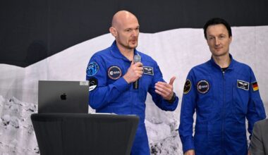 Two astronauts in blue flight suits talk into microphones at a lectern