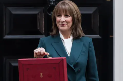 Getty Images Chancellor Rachel Reeves, who has shoulder-length brown hair, holds her red Budget briefcase with the black door of Number 11 Downing Street in the background. She is wearing a white blouse and a green trouser suit.