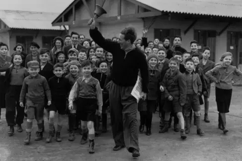 Fox Photos via Getty Images Black and white photo of a camp leader ringing the dinner bell at a camp for young Jewish 'Kindertransport', refugees from Germany and Austria. He is being followed by a large group of children