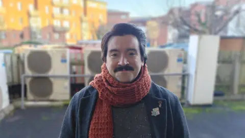 JR McCormick A man with brown hair and a moustache smiles at the camera. He is wearing a jumper and a jacket, along with a red scarf. He is standing outside. There are red-brick buildings in the background. 