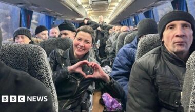Belarusian protest leader Maria Kolesnikova smiles and makes a heart-shaped gesture with her hands, on a bus full of former prisoners after their release. Maria is a woman with short brown hair, in a black jacket. The other prisoners we can see are men, many of them in black wool hats and winter coats.