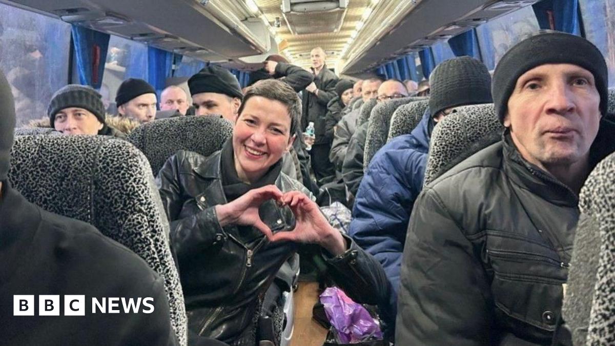 Belarusian protest leader Maria Kolesnikova smiles and makes a heart-shaped gesture with her hands, on a bus full of former prisoners after their release. Maria is a woman with short brown hair, in a black jacket. The other prisoners we can see are men, many of them in black wool hats and winter coats.