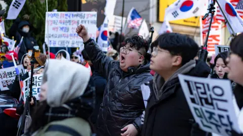 BBC/Hosu Lee A man in glasses and a black puffer jacket punches the air while yelling, surrounded by a crowd of people holding banners and South Korean flags