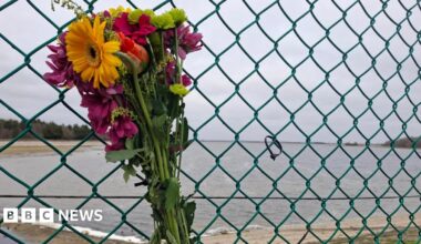 A floral bouquet that has been strapped to a metal fence. It is made of flowers that are different shades of yellow, orange, pink and red. A reservoir can be seen on the other side of the fence. It is a grey cloudy day.
