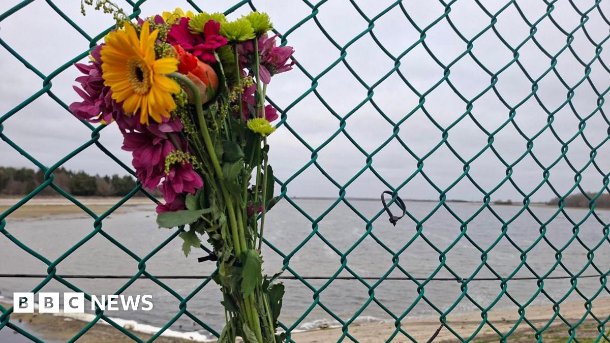 A floral bouquet that has been strapped to a metal fence. It is made of flowers that are different shades of yellow, orange, pink and red. A reservoir can be seen on the other side of the fence. It is a grey cloudy day.