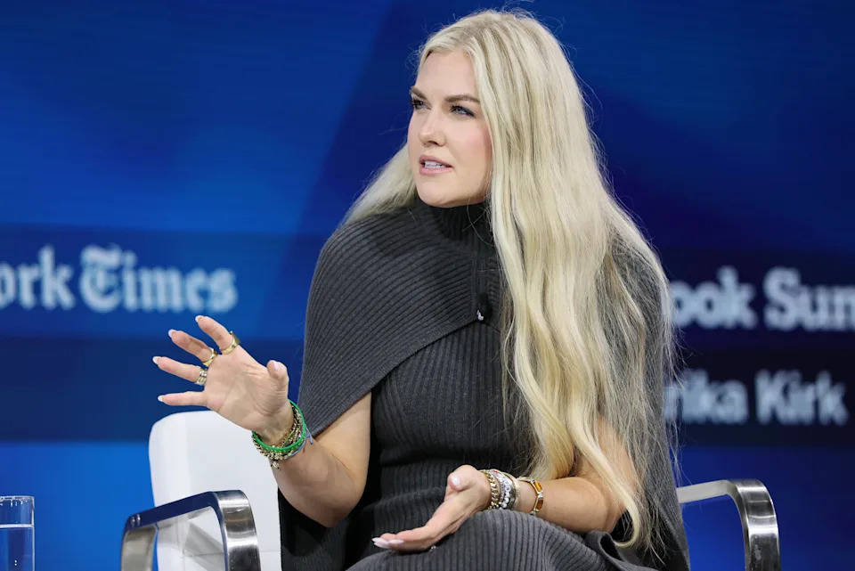 Person seated, speaking at an event, wearing a knitted turtleneck cape. The background shows a blurred event logo