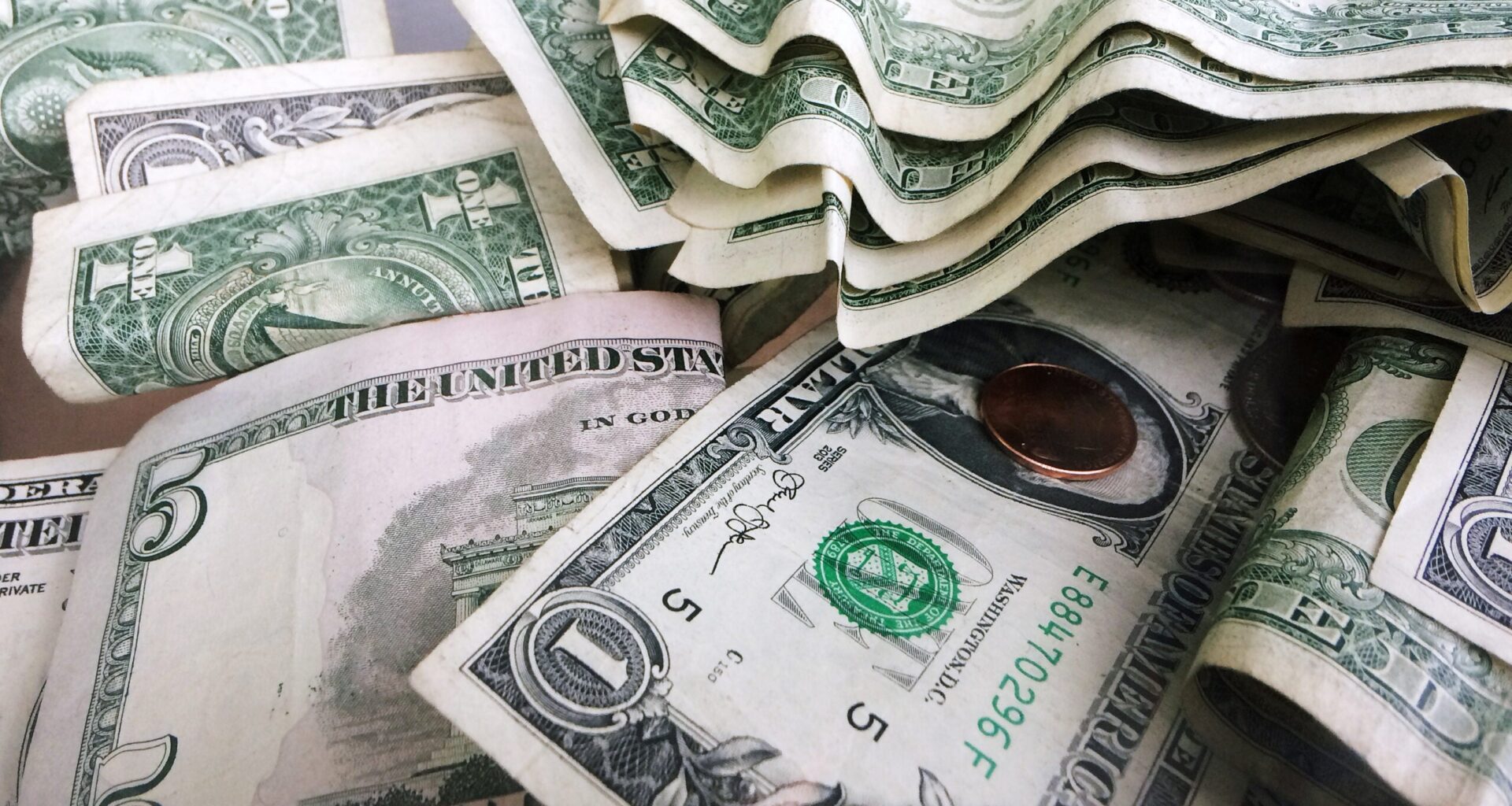 A close-up of assorted U.S. dollar bills and coins, including one- and five-dollar notes, scattered and partially stacked.