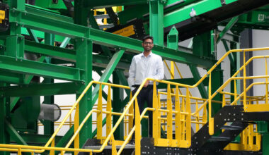 A man stands at the top of a yellow stairwell with his hand on the railing inside an industrial plant.